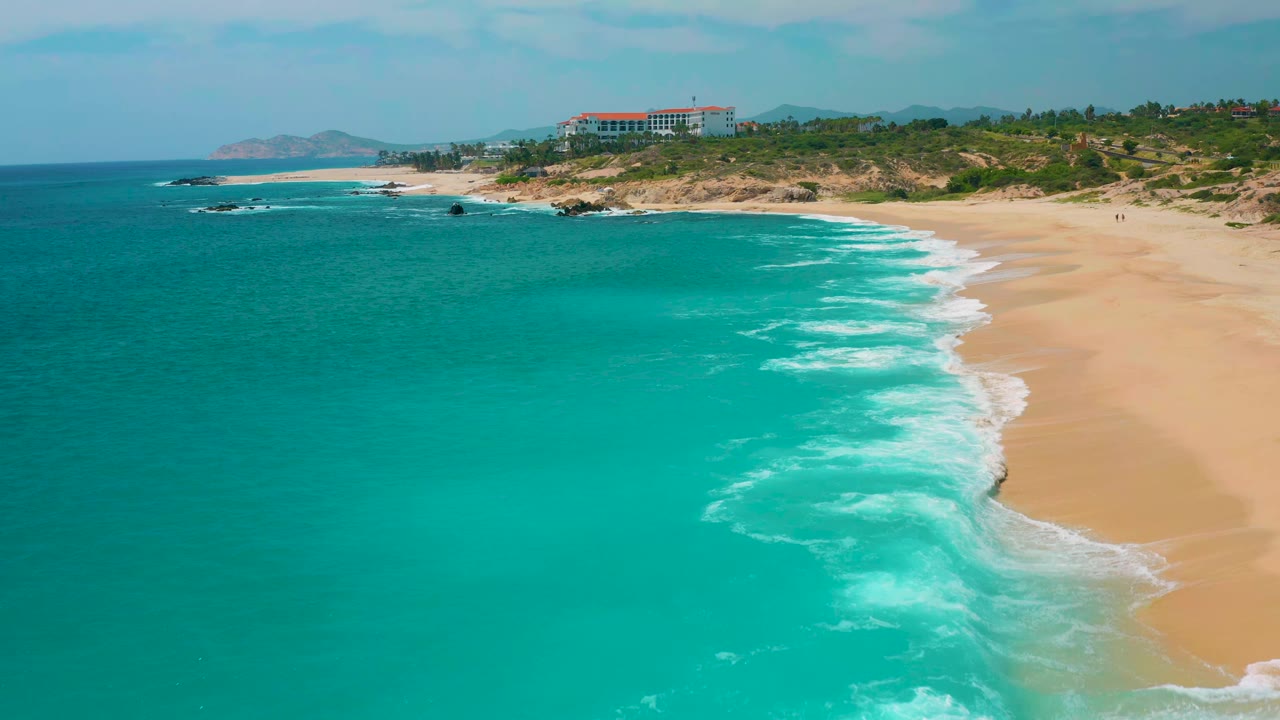 Drone view of blue-green water and diagonal surf lines along a broad beach.