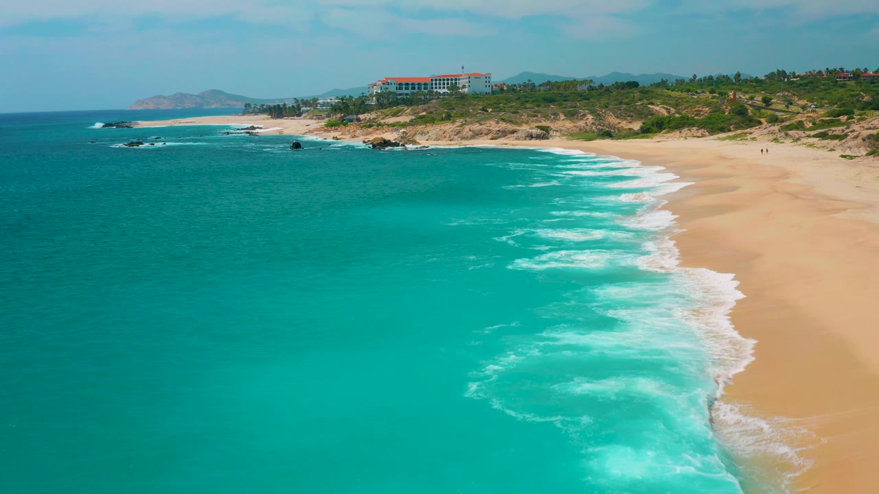 Aerial view at Secrets Marquis Los Cabos, Mexico, with turquoise water dominating the foreground, white foam curling toward the right edge of the beach, scrubby dunes beyond, and no visible people.