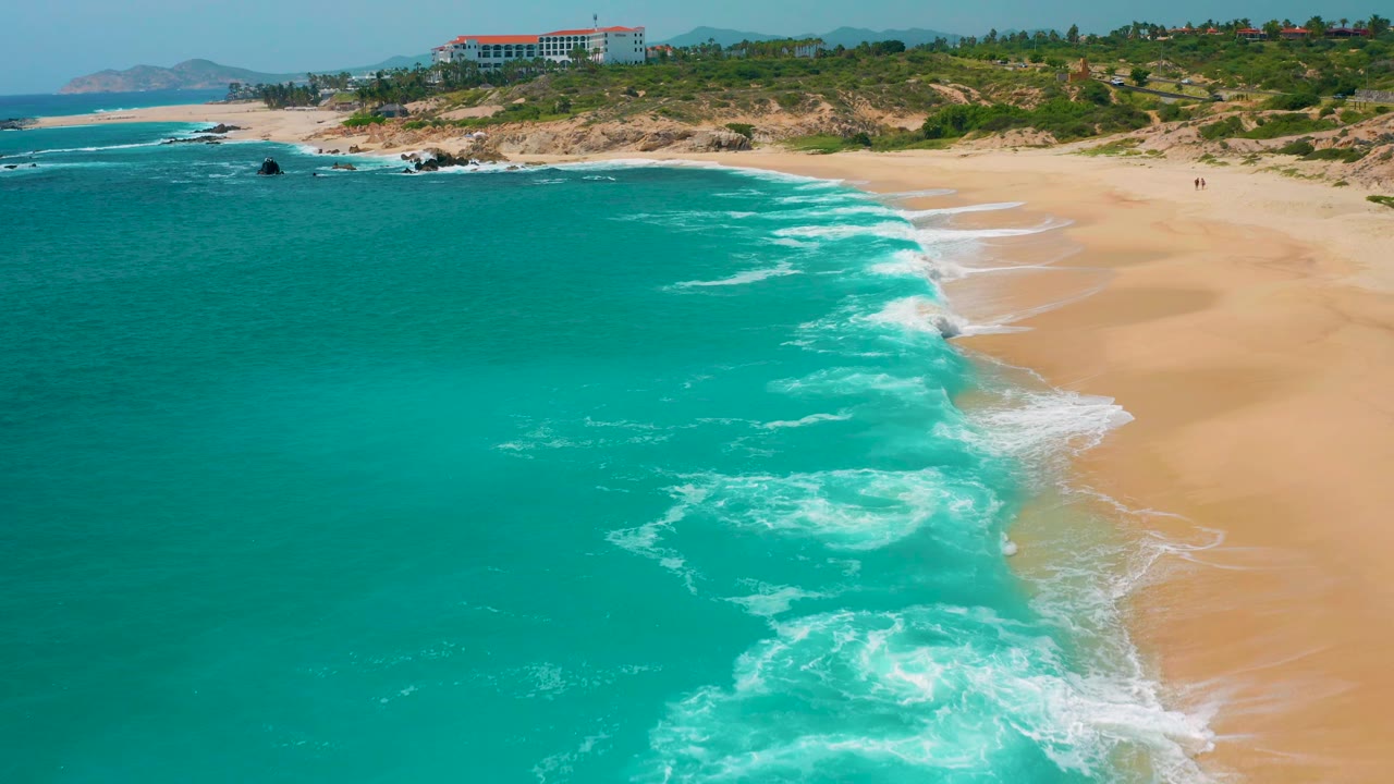 Aerial view over a Mexico beach with saturated turquoise water, white foam breaking diagonally onto pale sand, scrub-covered dunes behind the shore, and no clearly visible people.