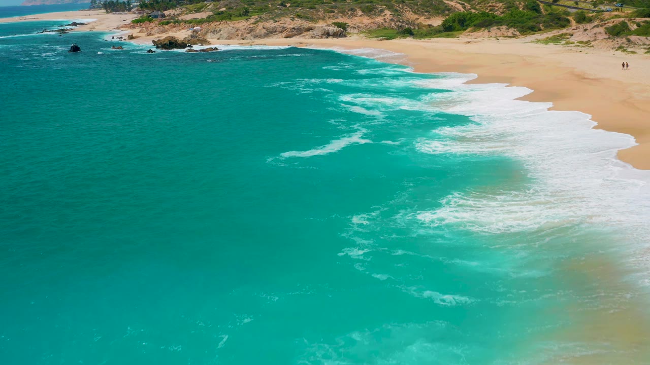 High aerial view over the Mexico shoreline with the ocean dominating the frame, a curving line of white foam near the sand, rocks along the upper coast, and no visible people.