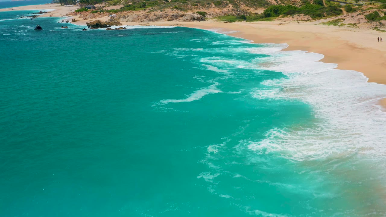 Aerial frame from the Mexico beach clip with clear blue-green water, a wide white surf wash on the right, sand beyond it, rocky coast near the top, and no visible people.