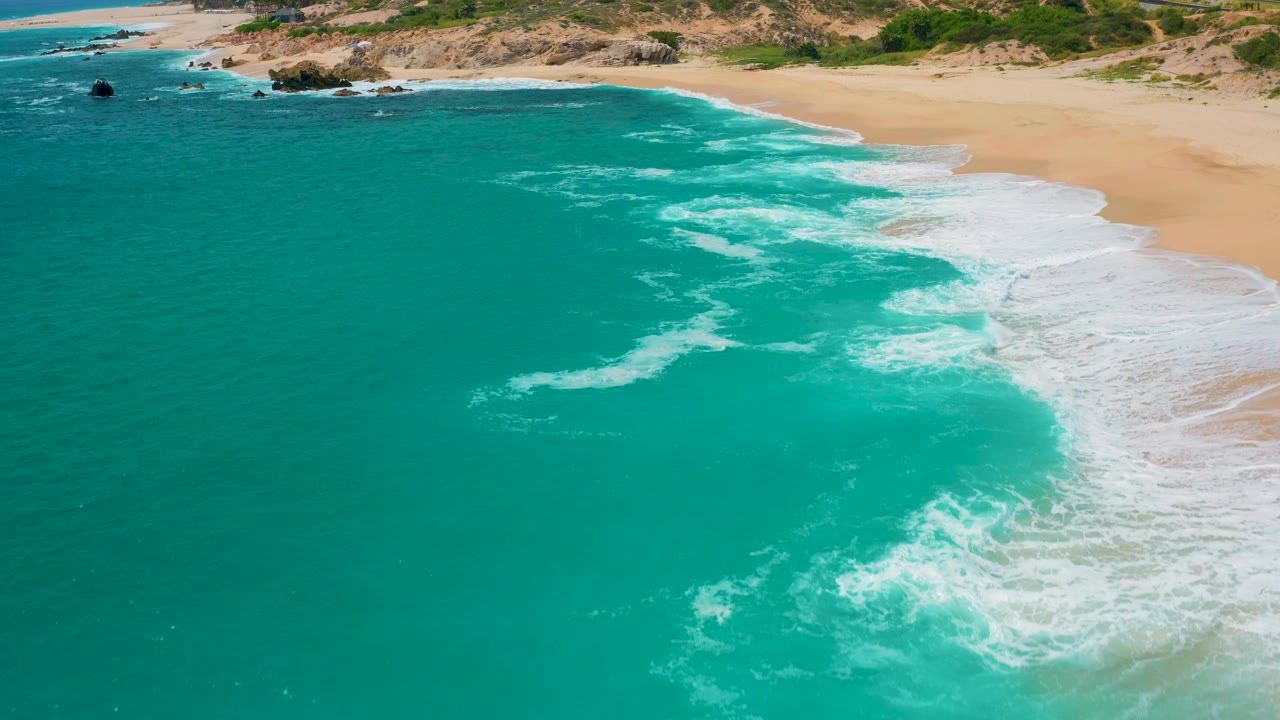 High aerial look at turquoise water in Mexico with thin white foam trails across the center, a sandy beach on the right edge, rocky shoreline above, and no visible people.