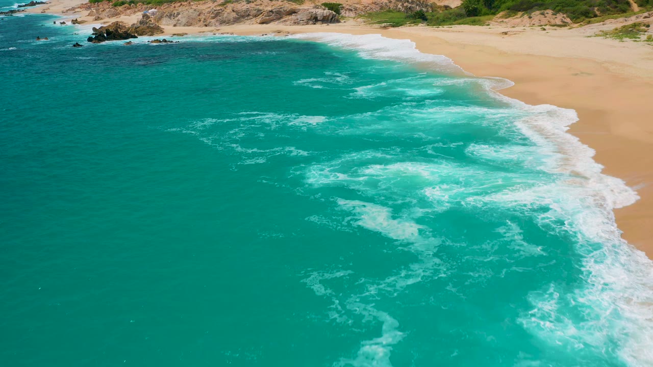 At 0:01, the aerial frame holds on clear blue-green water, loose white foam patches, a diagonal sandy shoreline, and no visible people at Secrets Marquis Los Cabos, Mexico.