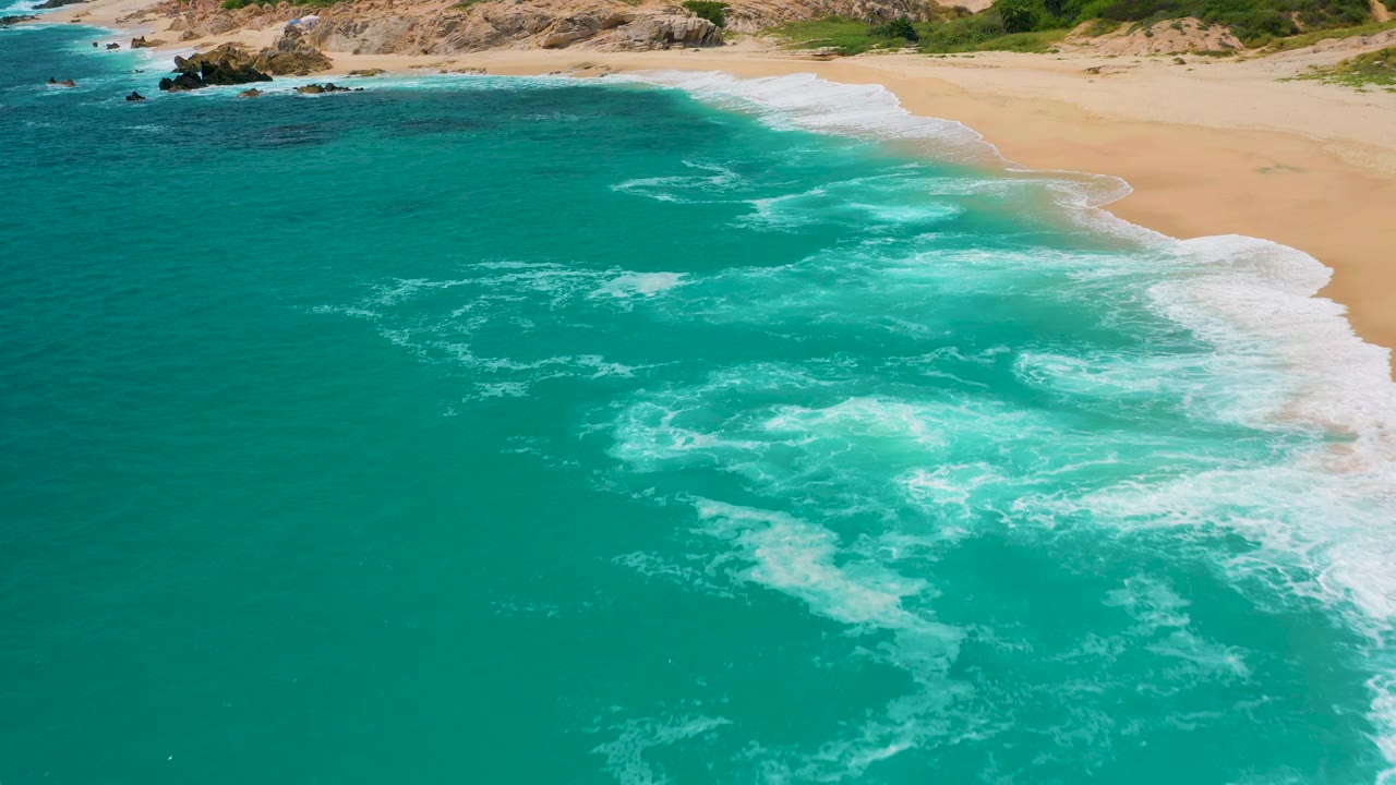 The 0:02 frame shows a wider patch of foamy turquoise surf pushing toward open golden sand, with rocks near the upper shoreline and no visible people.