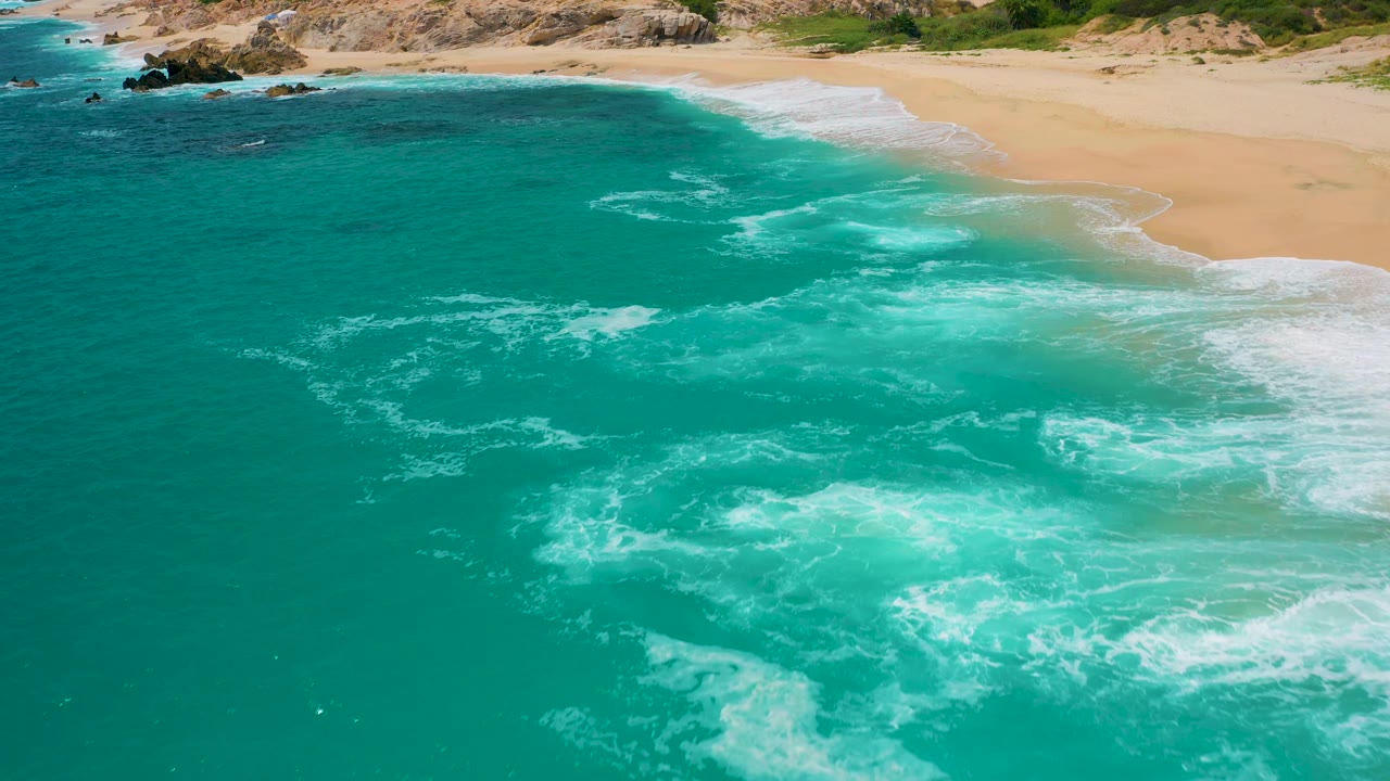 At 0:03, white foam spreads across the lower right water while the sandy beach arcs across the upper frame, with no visible people on the shore.