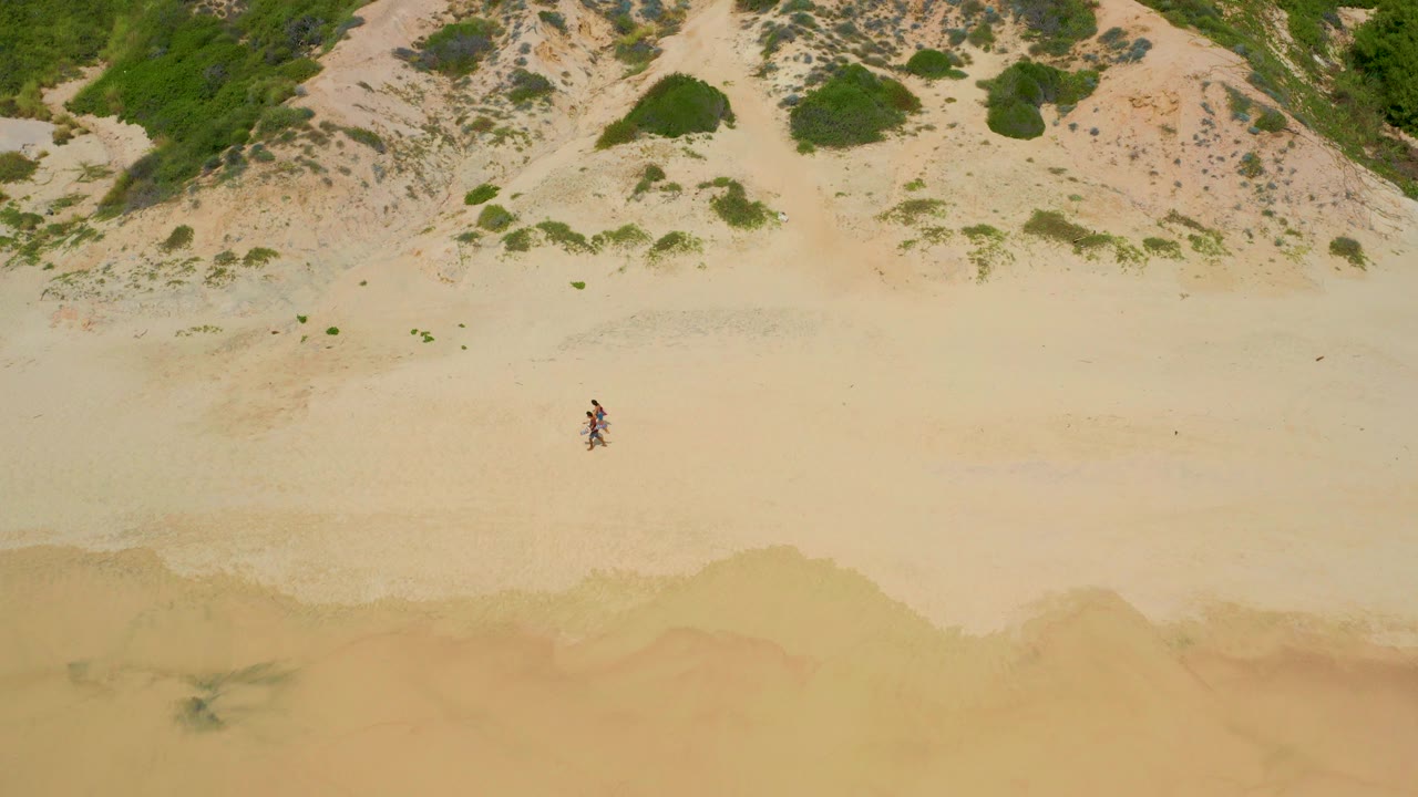 Overhead frame at Secrets Marquis Los Cabos, Mexico showing two small people on pale sand, dune vegetation above them, and wet shore wash near the lower edge.