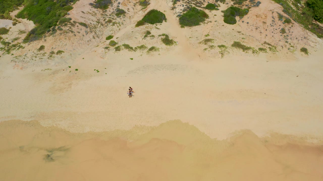Secrets Marquis Los Cabos, Mexico beach seen from above, with the walkers near the middle distance, textured dunes at the top, and soft tan water staining along the bottom.