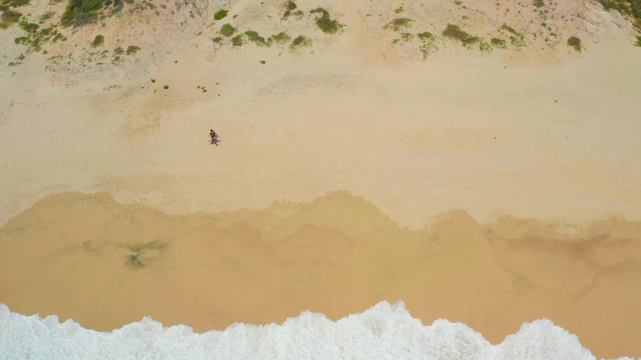 Overhead frame from Secrets Marquis Los Cabos, Mexico showing one small person walking on pale sand, dune vegetation along the top edge, and white surf entering from below.