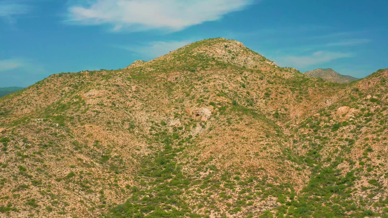 Rocky hillside fills the center near Secrets Marquis Los Cabos, Mexico, with patchy green vegetation, a pale cloud above, distant ridges, and no visible people.