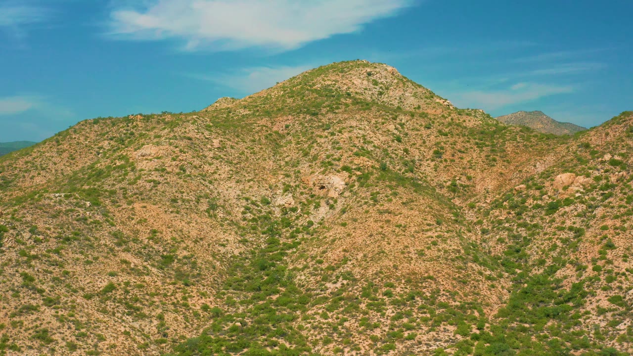 A sunlit desert mountain ridge near Secrets Marquis Los Cabos, Mexico, shows tan rock, scattered shrubs, small valleys, blue sky, and no visible people.