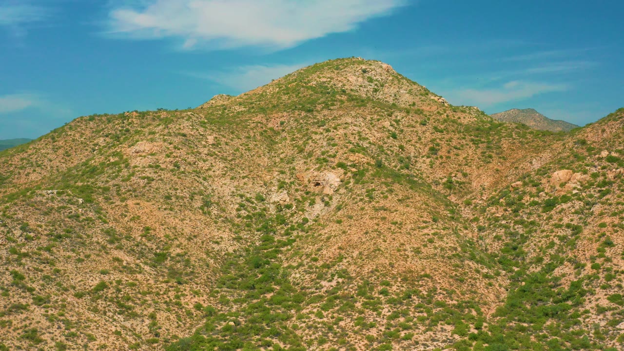 Arid hills near Secrets Marquis Los Cabos, Mexico, stretch across the frame with green washes in shallow valleys, tan rocky ground, blue sky, and no visible people.
