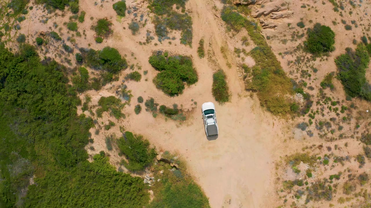 A top-down view in Secrets Marquis Los Cabos, Mexico shows the white truck slightly lower in frame on pale sand, with green brush clusters and dry scrub around it, no visible people.