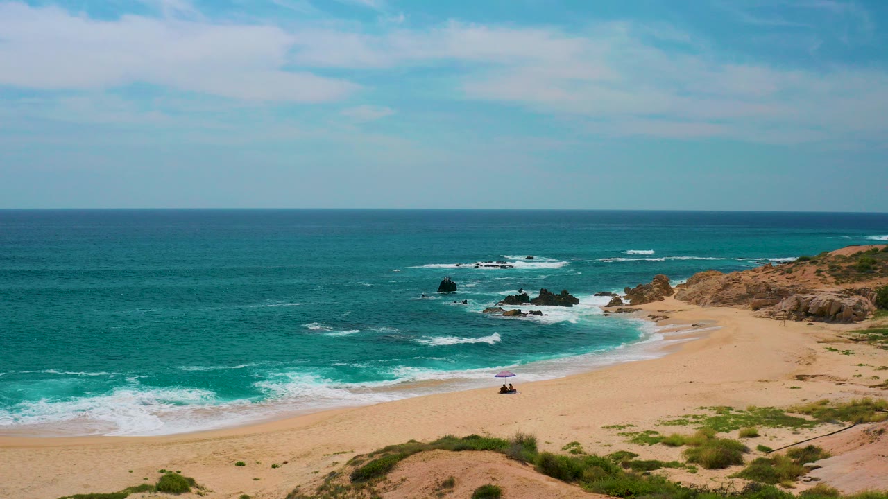 Opening view at Secrets Marquis Los Cabos in Mexico showing turquoise surf rolling onto a sandy beach beside dark rocks and coastal vegetation, with tiny beachgoers visible near the lower center.