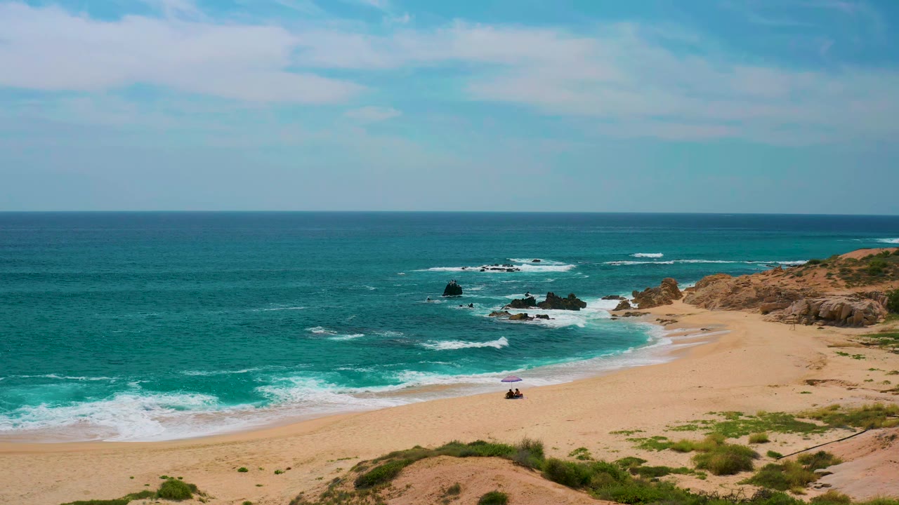 At 0:01, the Mexico shoreline remains wide in frame as white water spreads along the sand and the small purple umbrella and beachgoers sit below the rocky right side.