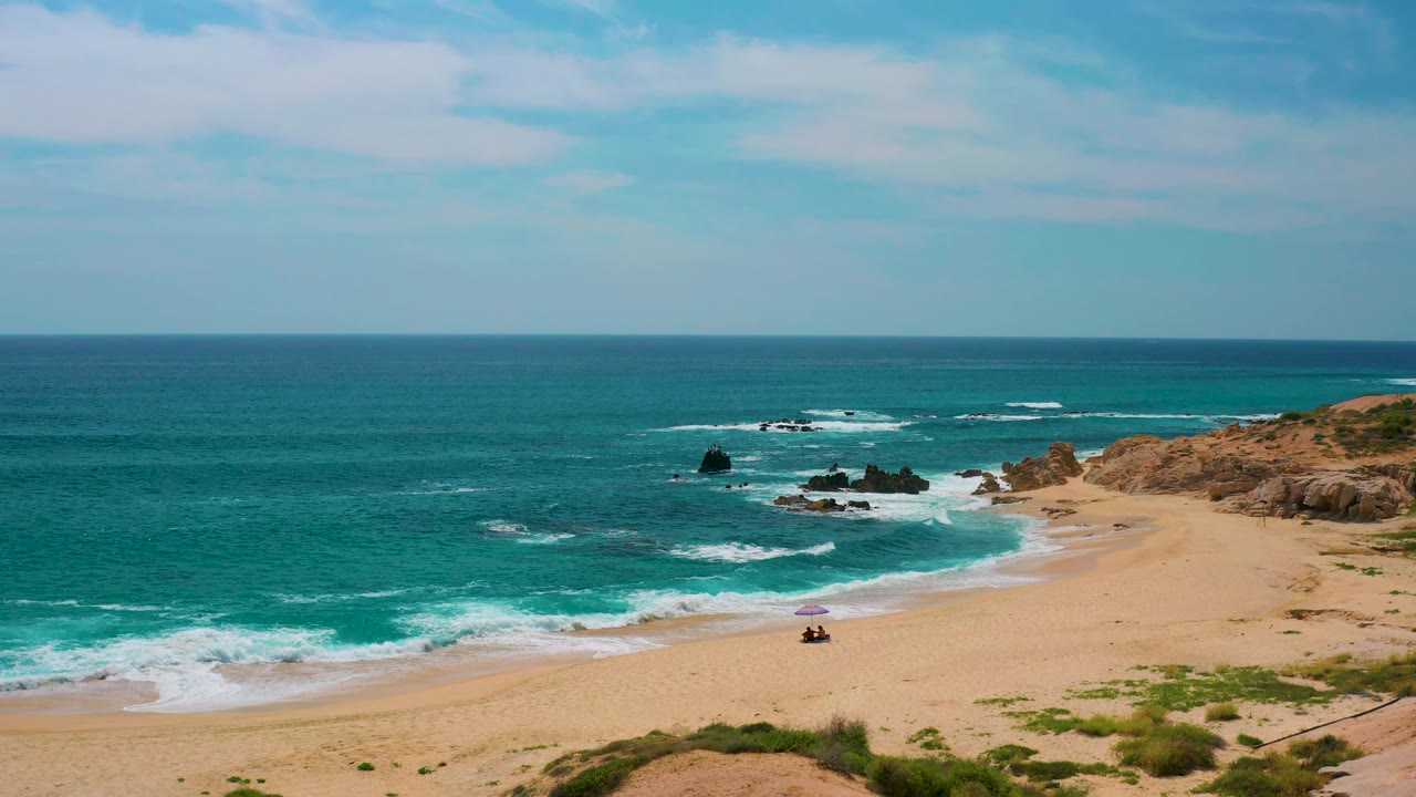 At 0:02, the Secrets Marquis Los Cabos beach curve is centered with surf wrapping toward the right-hand rocky point and tiny beachgoers visible as small dark shapes near a purple umbrella.