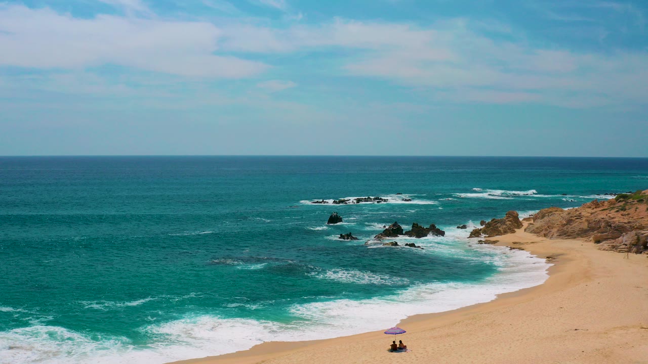 Aerial frame from Secrets Marquis Los Cabos, Mexico showing turquoise water, white surf, golden sand, dark rocks offshore, and no clearly visible people.