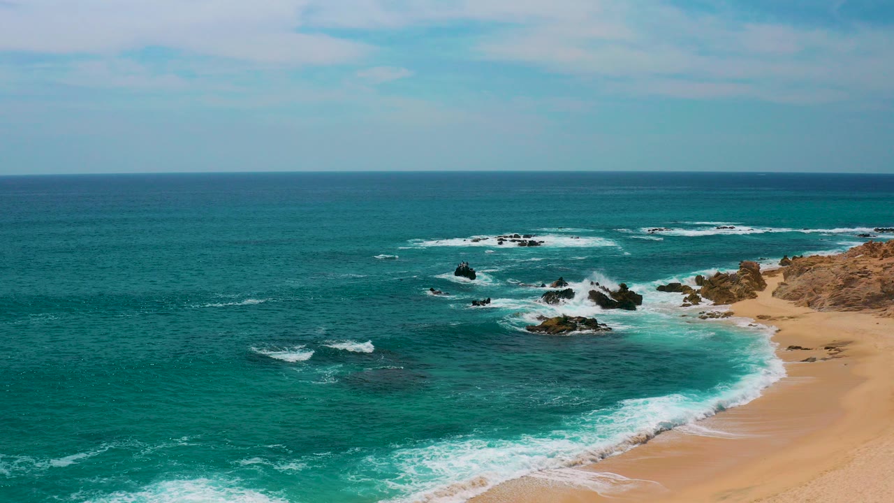 Aerial frame at Secrets Marquis Los Cabos, Mexico, showing teal ocean, tan sand, dark rock clusters, white foam at the shore, and no visible people.