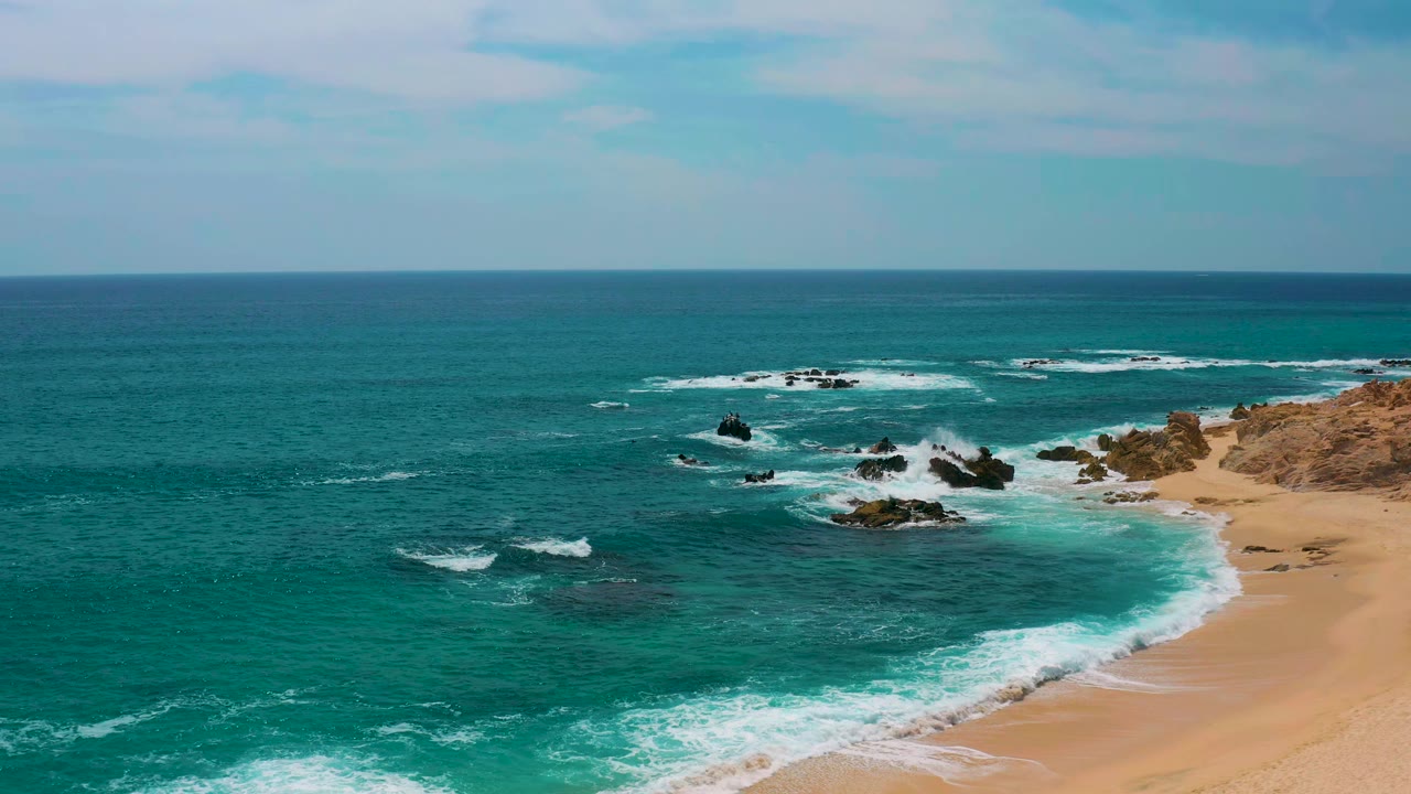 Aerial view at Secrets Marquis Los Cabos, Mexico, with a wider sweep of turquoise water, white breakers around offshore rocks, tan beach at right, and no visible people.