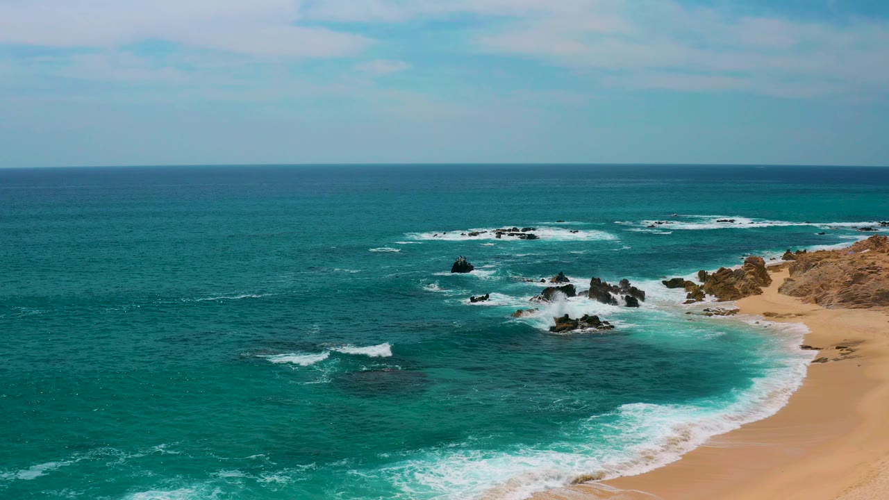 Aerial frame at Secrets Marquis Los Cabos, Mexico, showing more open water on the left, foamy surf lines moving toward the beach, scattered black rocks, and no visible people.
