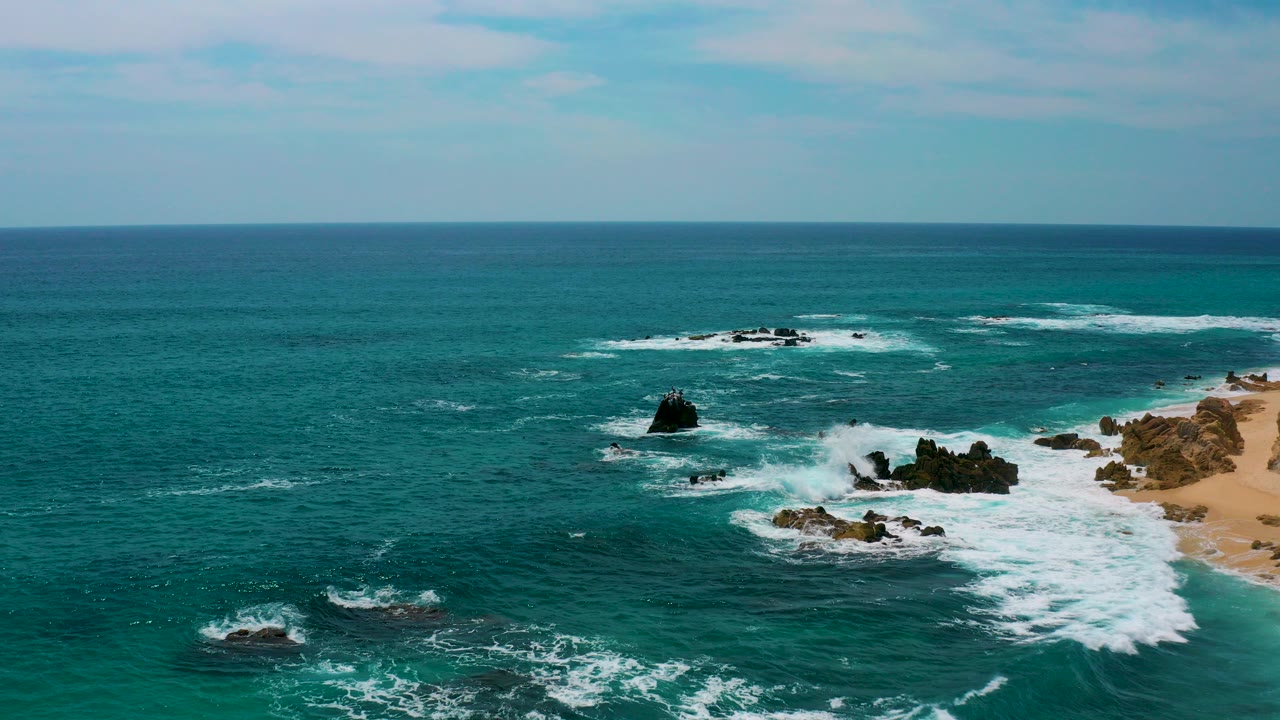 Aerial view of turquoise ocean, white surf, dark rocks, and a strip of sandy beach.