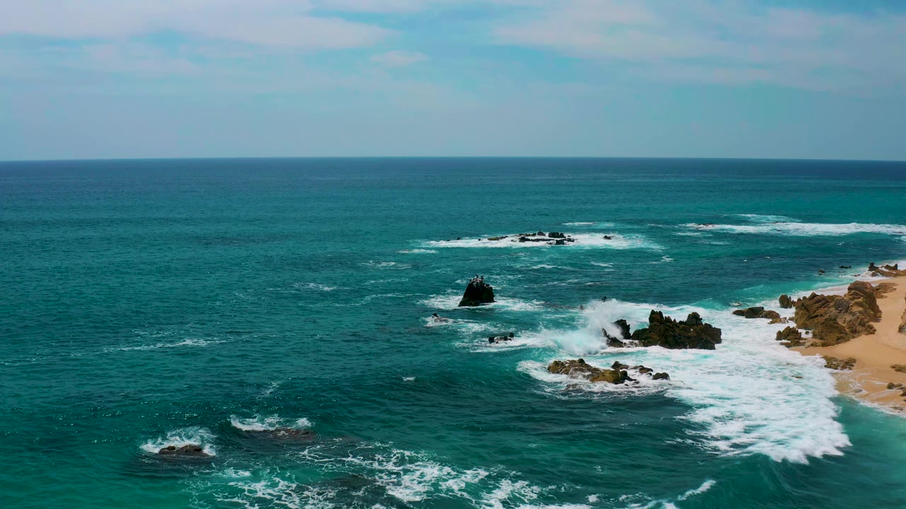 Aerial frame from Secrets Marquis Los Cabos, Mexico, with foamy waves wrapping around central black rocks, open teal water in front, sandy shoreline at right, and no visible people.