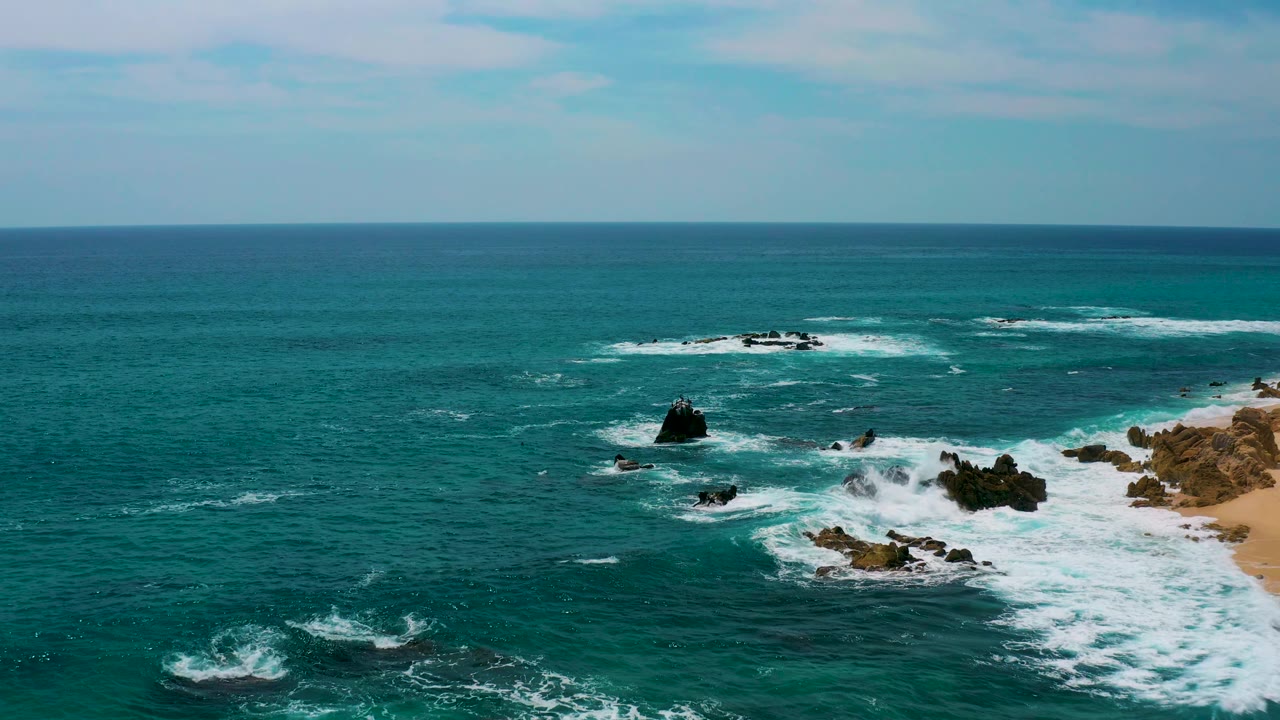 Wide aerial frame at Secrets Marquis Los Cabos, Mexico, showing scattered dark rocks, broad turquoise water, breaking white surf, partial sand on the right edge, and no visible people.