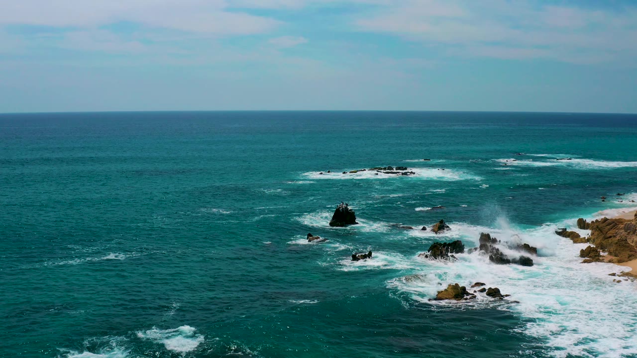 Elevated frame from Secrets Marquis Los Cabos, Mexico, with a tall dark rock left of center, white water over low rocks, open ocean behind, a thin beach edge at right, and no visible people.