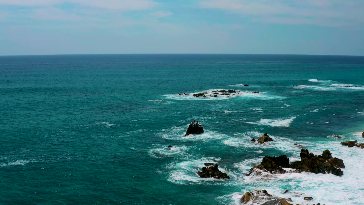 Wide aerial frame at Secrets Marquis Los Cabos, Mexico, showing a central dark rock, low reef-like rocks near the horizon, white wave streaks, and no visible people.
