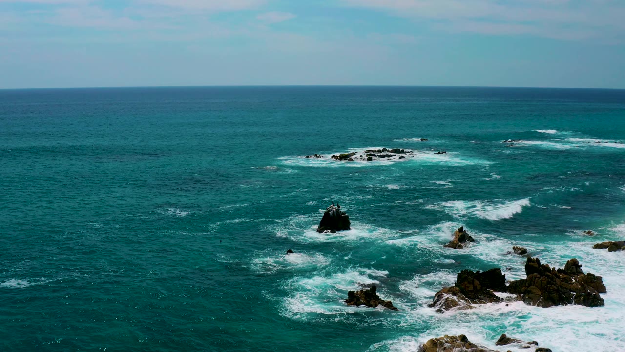 Aerial coastal frame at Secrets Marquis Los Cabos, Mexico, with teal water, a black rock stack left of center, foamy water near the lower right rocks, and no visible people.