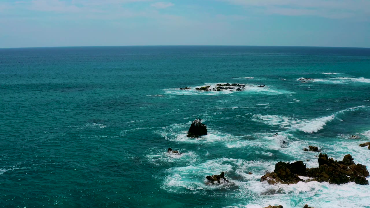 Elevated view at Secrets Marquis Los Cabos, Mexico, with blue-green water, isolated dark rocks, broad white foam patches, a pale sky, and no visible people.
