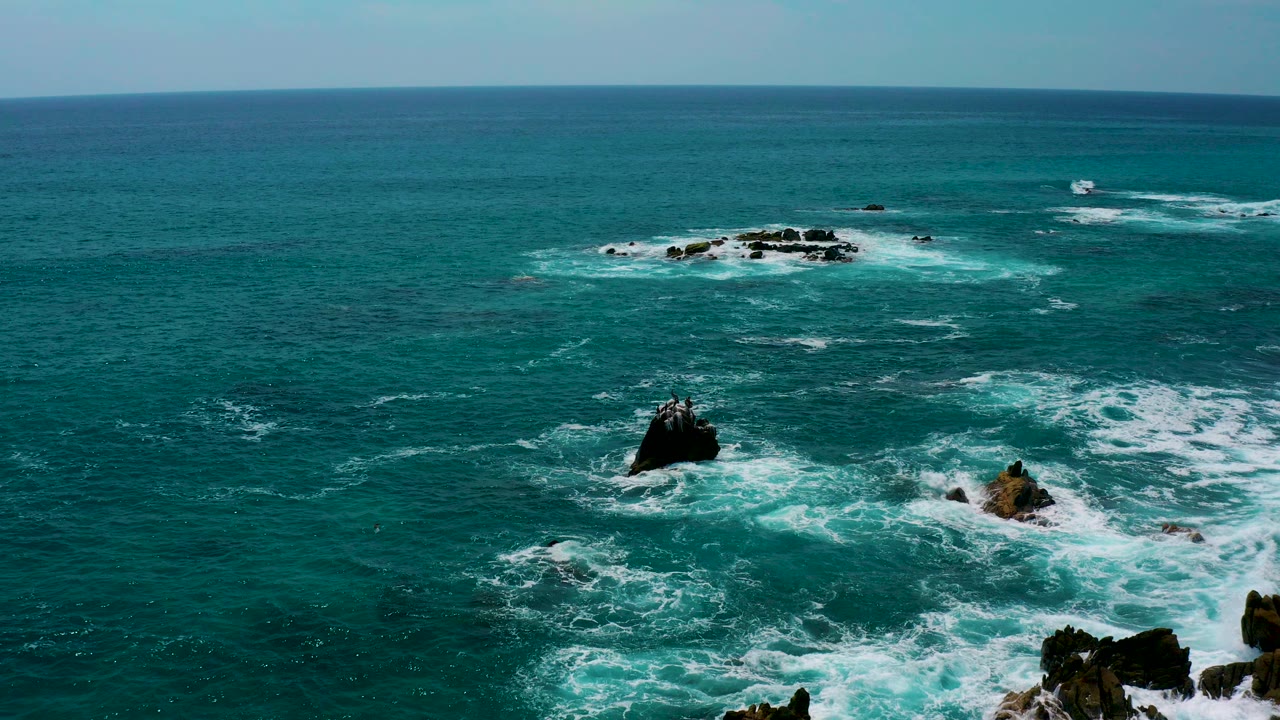 Aerial view over blue-green ocean at Los Cabos, Mexico, with dark rocks in the lower frame, white surf around them, a smaller rock cluster near the horizon, and no visible people.