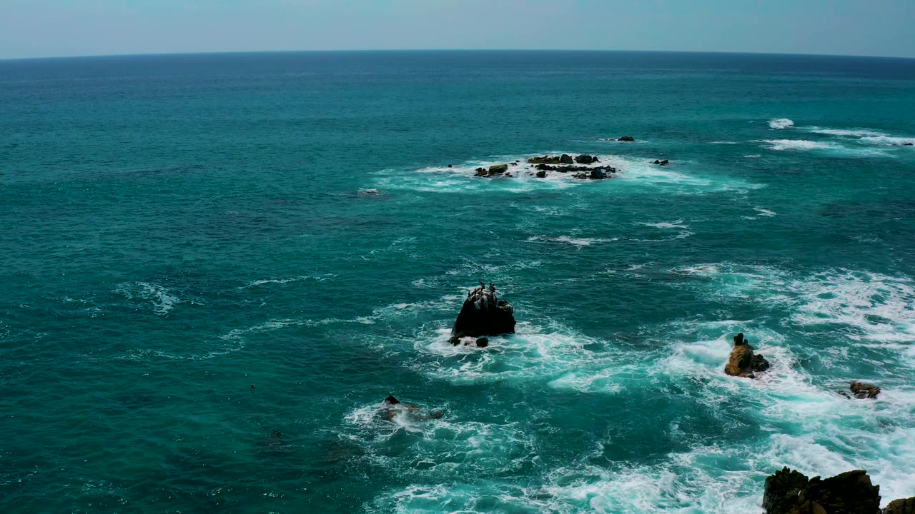 Aerial frame from Los Cabos, Mexico, showing open blue water, a dark rock near the lower center, a tan rock to the right, foam trails between formations, and no visible people.