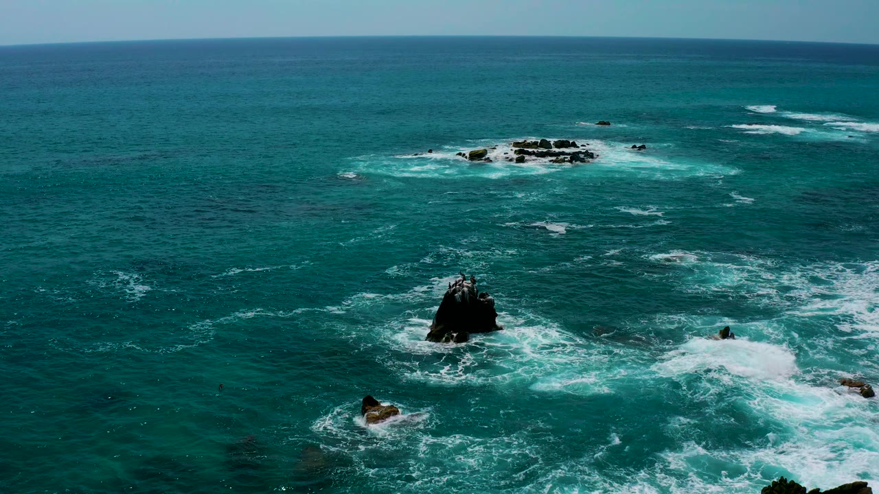 Wide elevated view of the Los Cabos coast water with a dark rock in the lower middle, a bright patch of breaking surf to the right, distant rocks near the horizon, and no visible people.