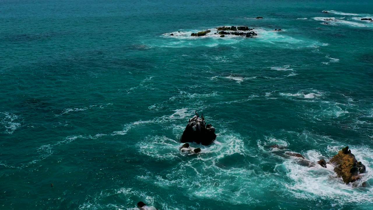 Aerial ocean frame near Secrets Marquis Los Cabos, Mexico, showing a dark rock in the lower center, a brown rock to the right, and broken foam lines across open water with no visible people.