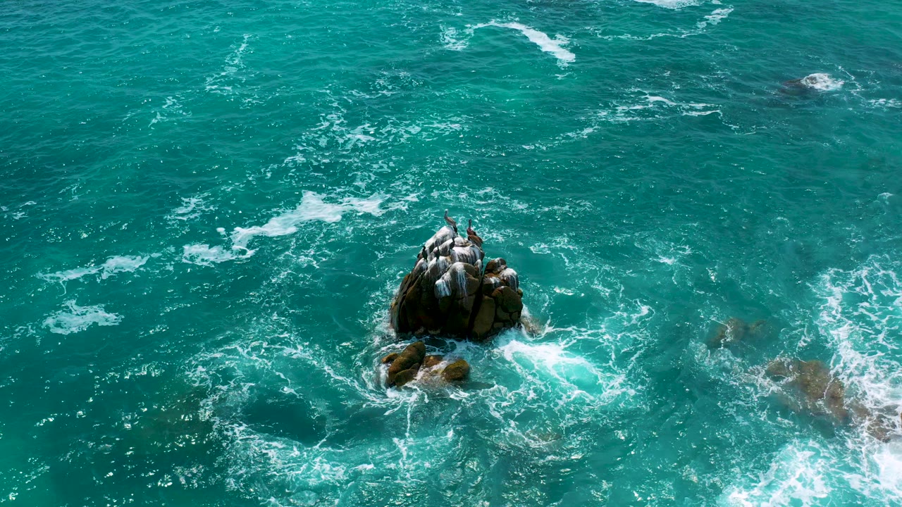 Aerial view over Secrets Marquis Los Cabos, Mexico, with a dark rock outcrop surrounded by turquoise water and white foam, no visible people.