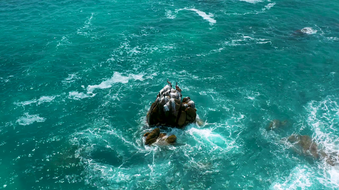 Aerial frame over Secrets Marquis Los Cabos, Mexico, showing the rock formation slightly lower in frame with whitewater curling around nearby submerged rocks, no visible people.