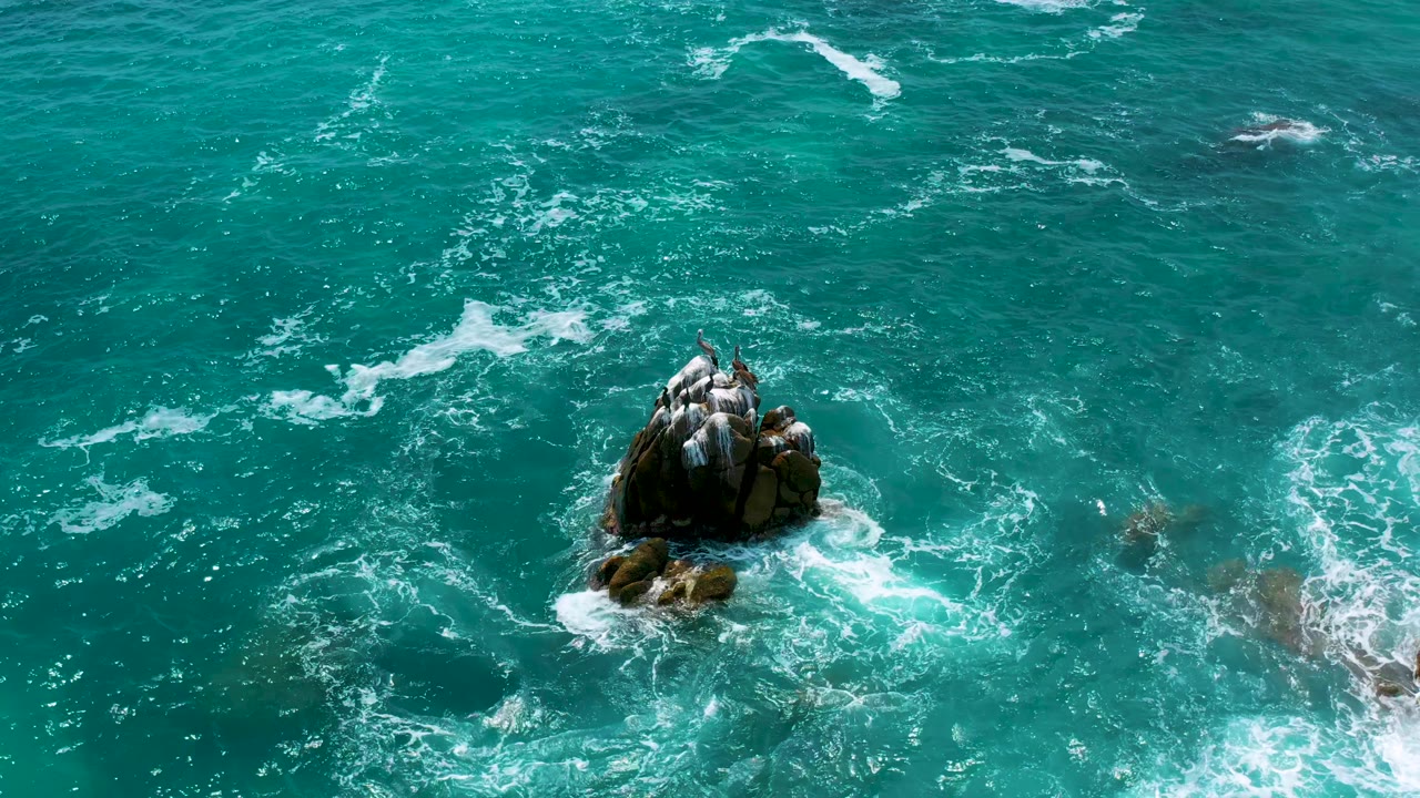 Aerial view at Secrets Marquis Los Cabos, Mexico, with the dark rock outcrop below center and a fresh patch of white surf forming to the right, no visible people.