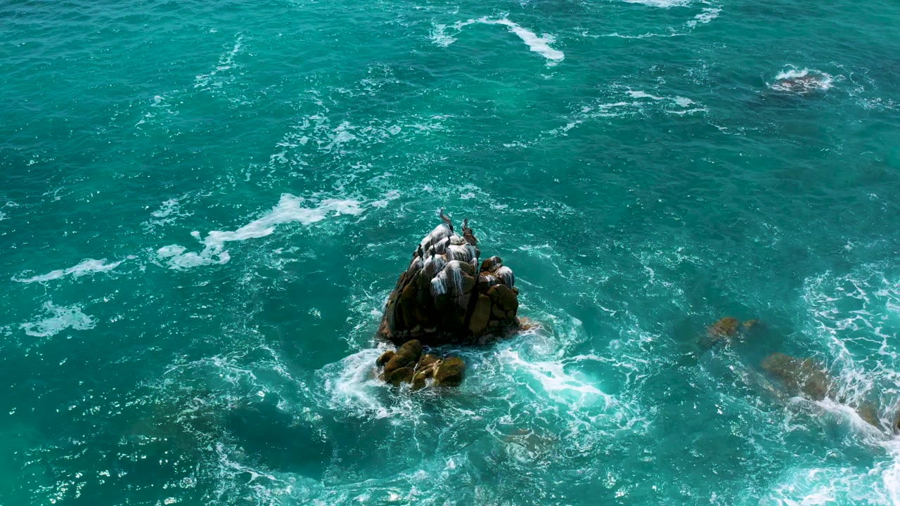 Top-down aerial frame over Secrets Marquis Los Cabos, Mexico, with the main rock low in the image and pale foam streaks crossing the surrounding water, no visible people.