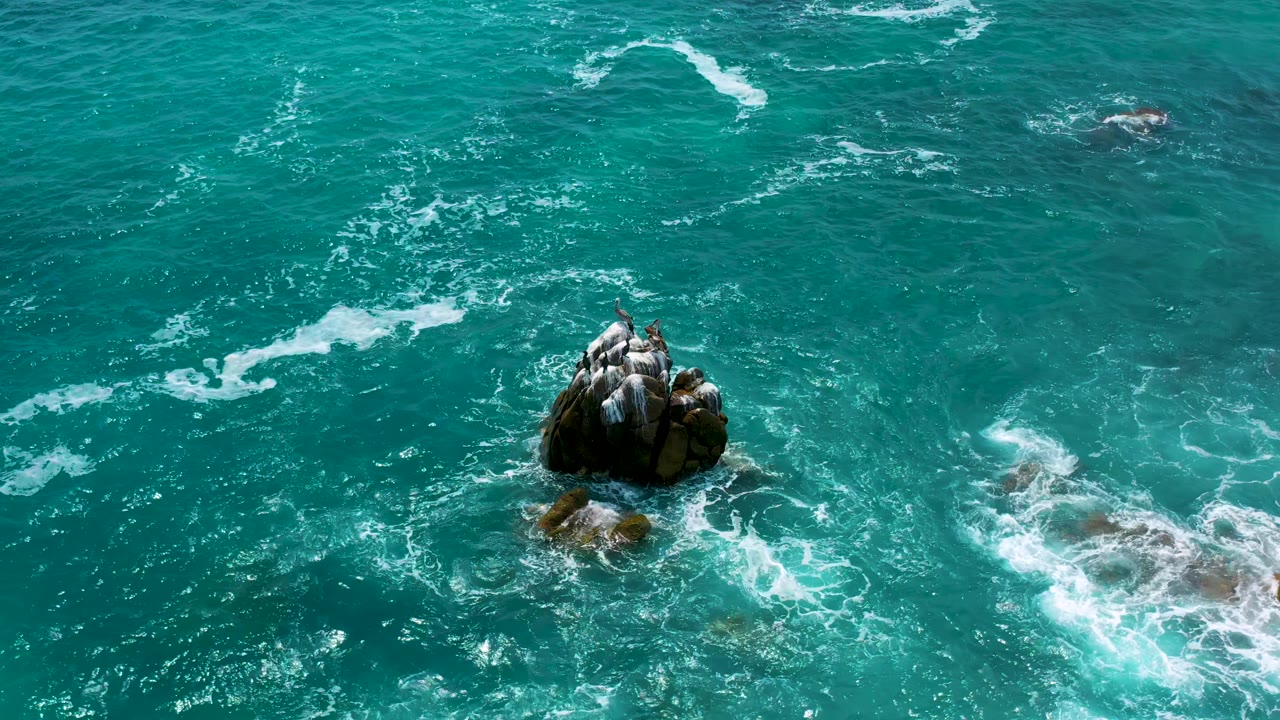 High-angle view at Secrets Marquis Los Cabos, Mexico, where a dark rock outcrop sits below center and bright foam breaks along rocks on the right, with no visible people.