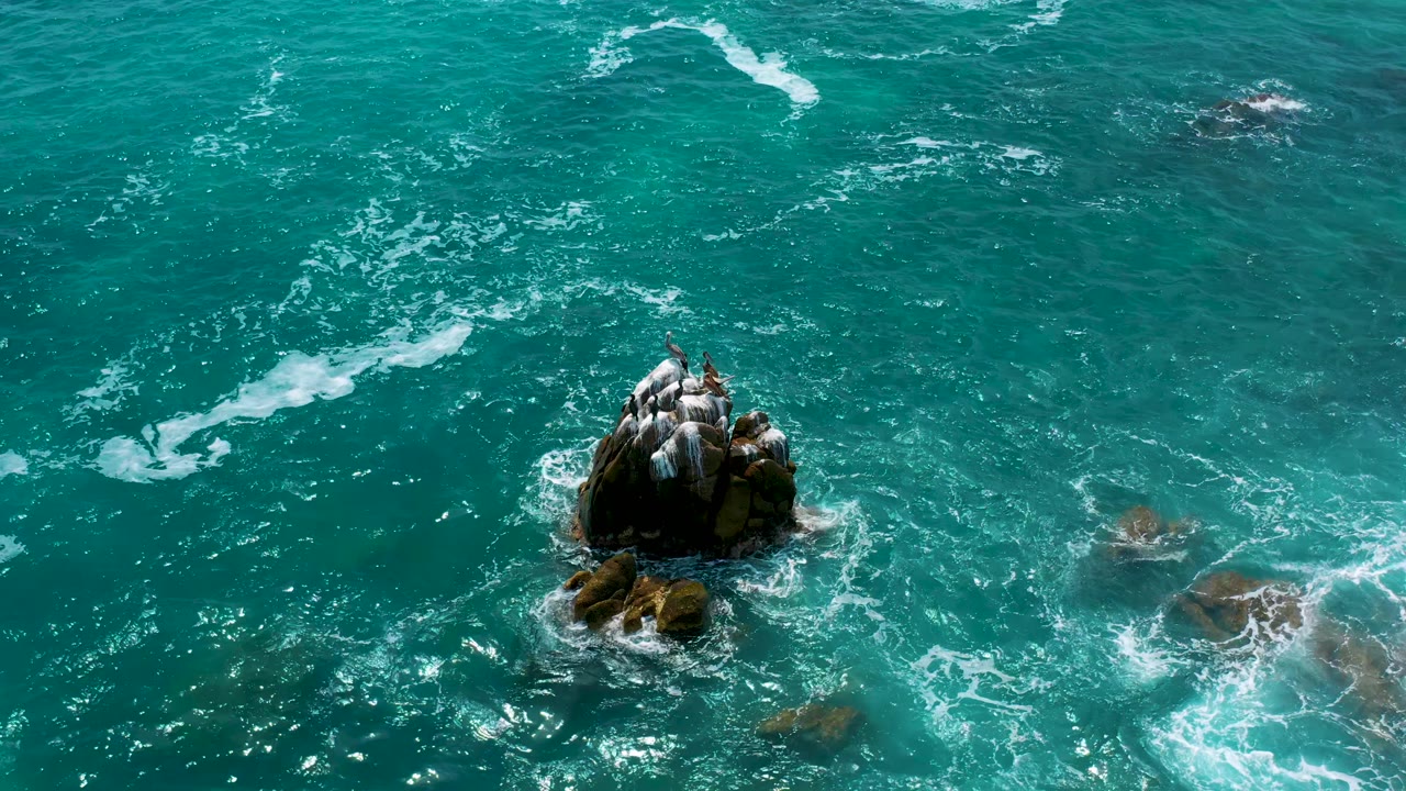 Aerial frame over Secrets Marquis Los Cabos, Mexico, showing the central rocks with foam streaks drifting across the surrounding blue sea and no visible people.
