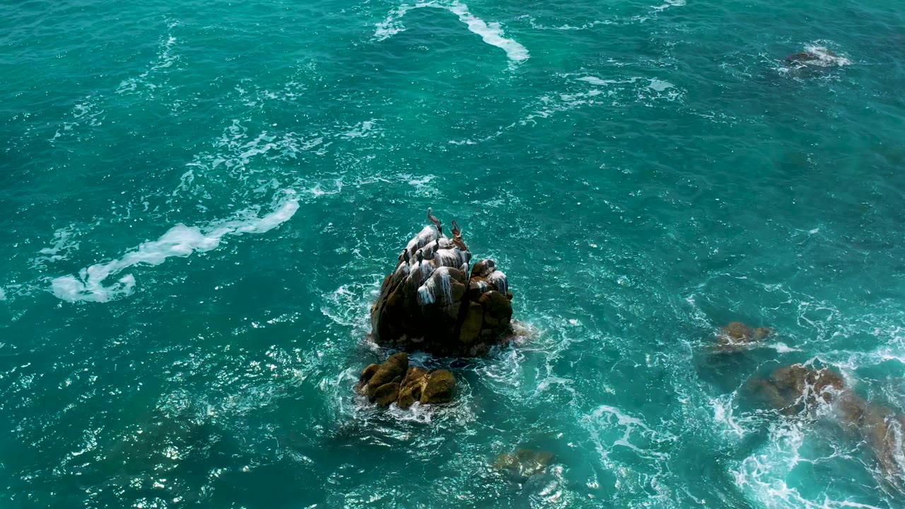 The overhead view holds on a dark rock outcrop at Secrets Marquis Los Cabos, Mexico, with pale surf marks and submerged rocks visible to the right, no visible people.