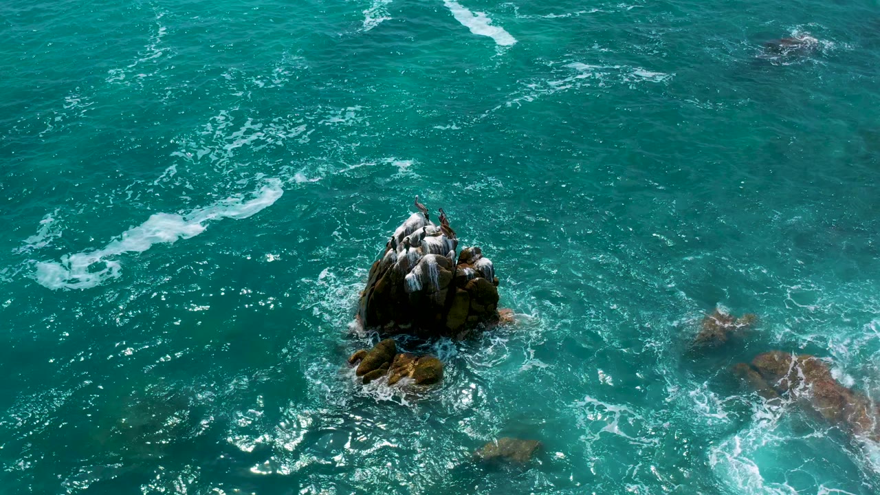 Overhead coastal water at Secrets Marquis Los Cabos, Mexico, with the main dark rocks centered low in frame, foam gathering below, and no visible people.