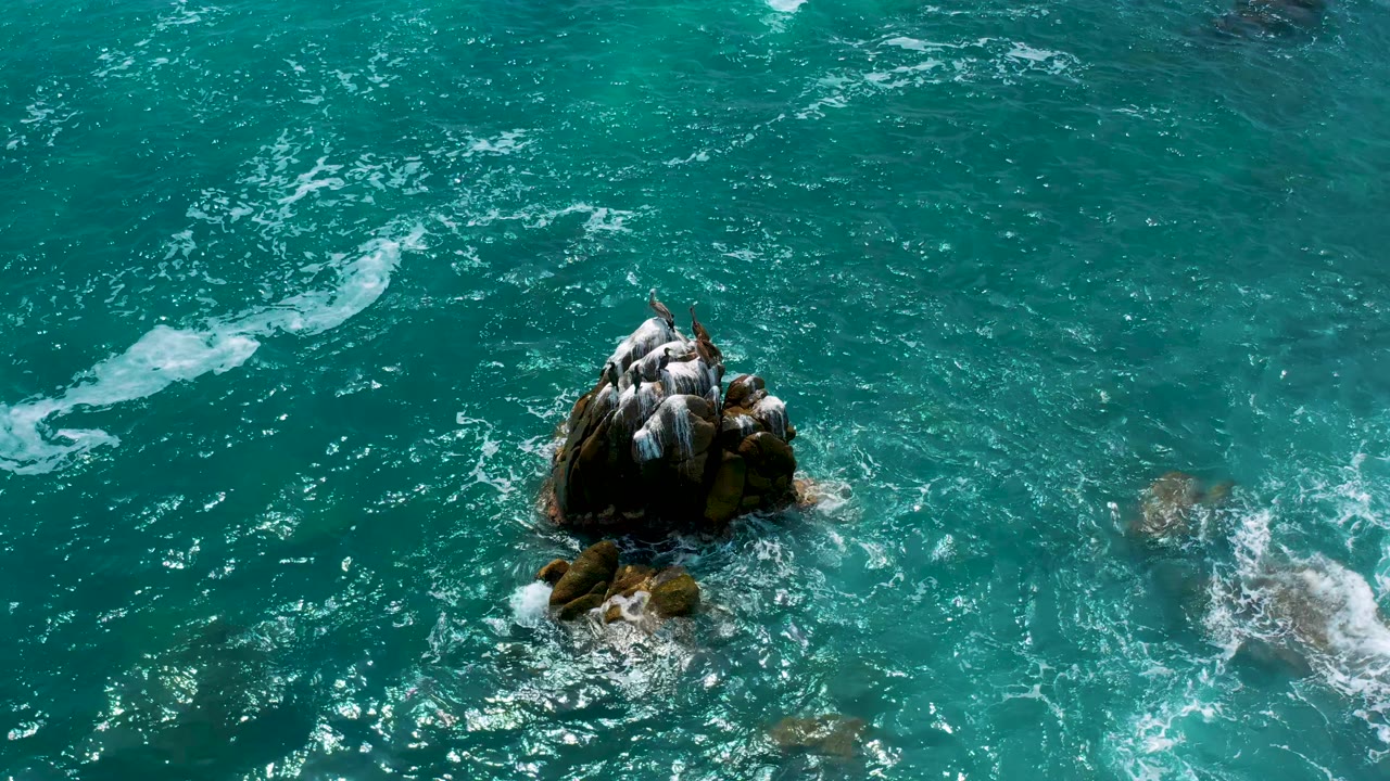 Aerial view of blue-green sea at Secrets Marquis Los Cabos, Mexico, with the main rock slightly left of center, foam streaks above it, and no visible people.