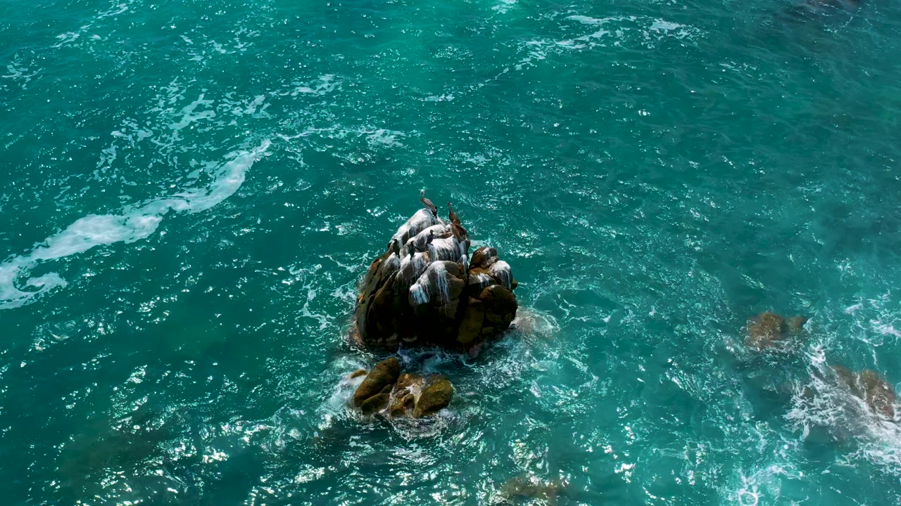 Top-down frame at Secrets Marquis Los Cabos, Mexico, showing the rocky outcrop lower in the frame, white surf curling at right, and no visible people.