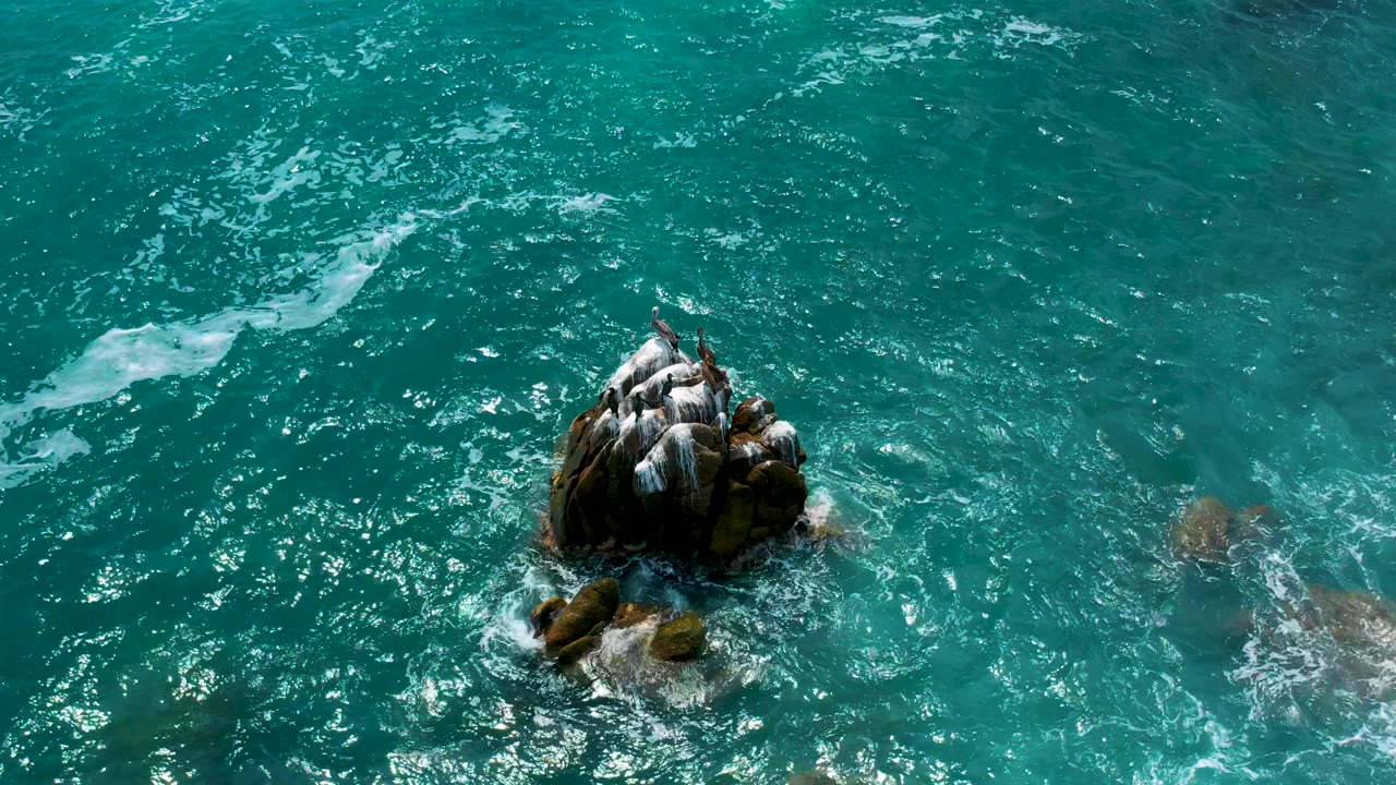 Overhead coastal water view at Secrets Marquis Los Cabos, Mexico, with the rock outcrop low and left, open turquoise water above, scattered foam, and no visible people.