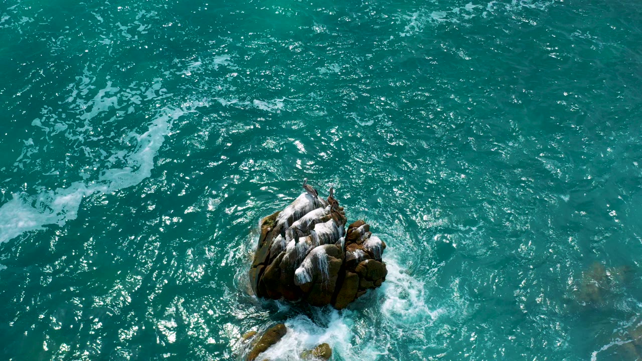 At Secrets Marquis Los Cabos, Mexico, an overhead view shows turquoise ocean water circling a dark rock outcrop with white foam at the lower edge and no visible people.
