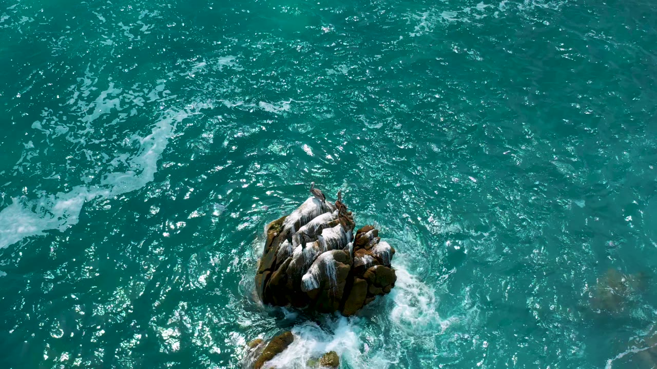At Secrets Marquis Los Cabos, Mexico, the rock remains near the center as bright ripples and small foam lines spread through the turquoise water, with no visible people.