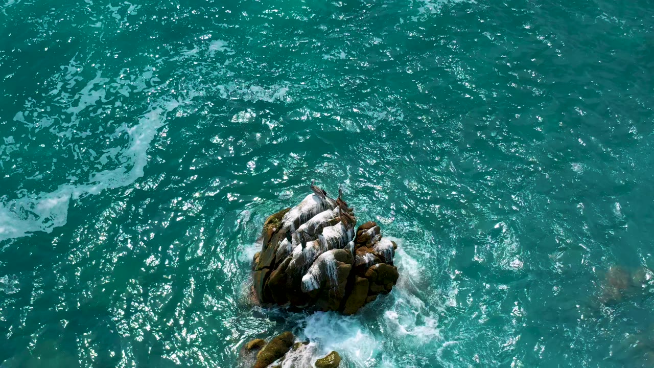 At Secrets Marquis Los Cabos, Mexico, white foam thickens on the lower side of the rock while clear teal water fills the surrounding frame, with no visible people.