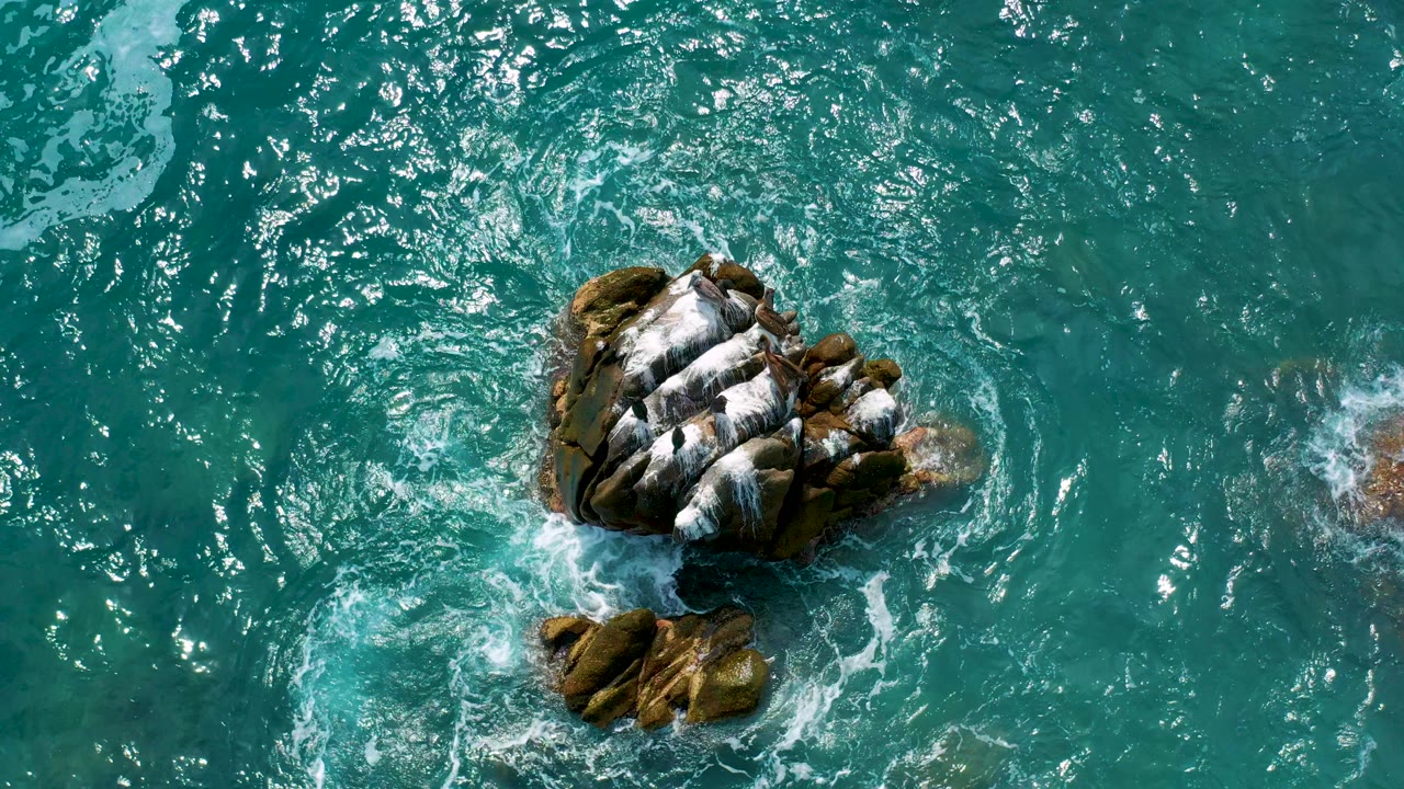 Overhead view at Secrets Marquis Los Cabos, Mexico, showing turquoise ocean swirling around a brown rock capped with white foam, with no visible people.