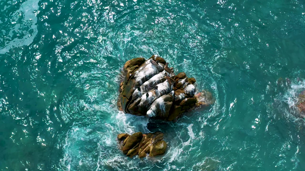 Overhead frame from Secrets Marquis Los Cabos, Mexico, with the rock centered slightly left and white foam streaking across its top while ripples fill the surrounding water, with no visible people.